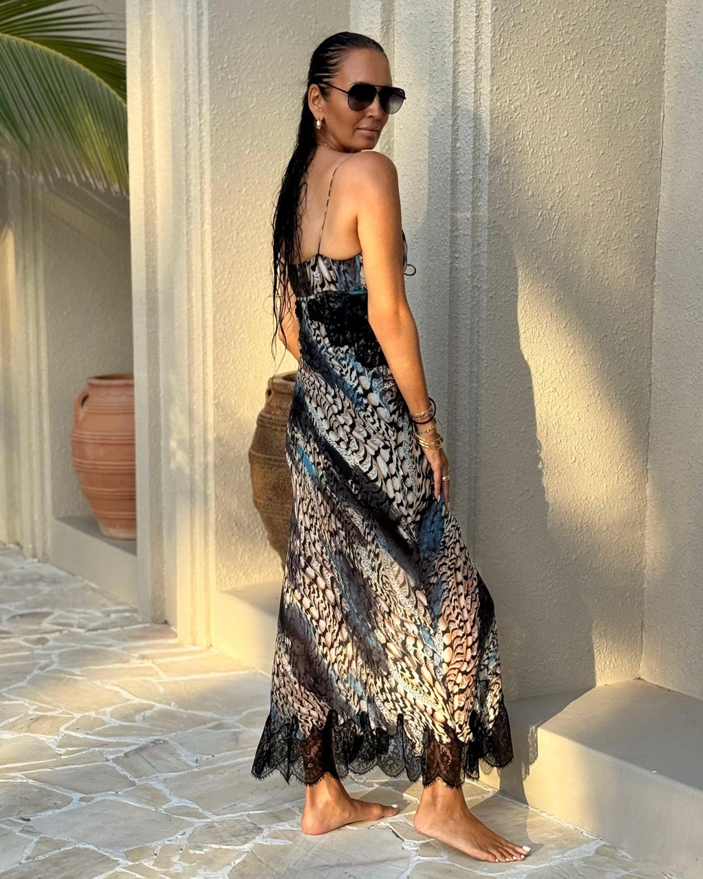 Woman in a patterned dress standing against a textured wall with sunlight casting shadows.
